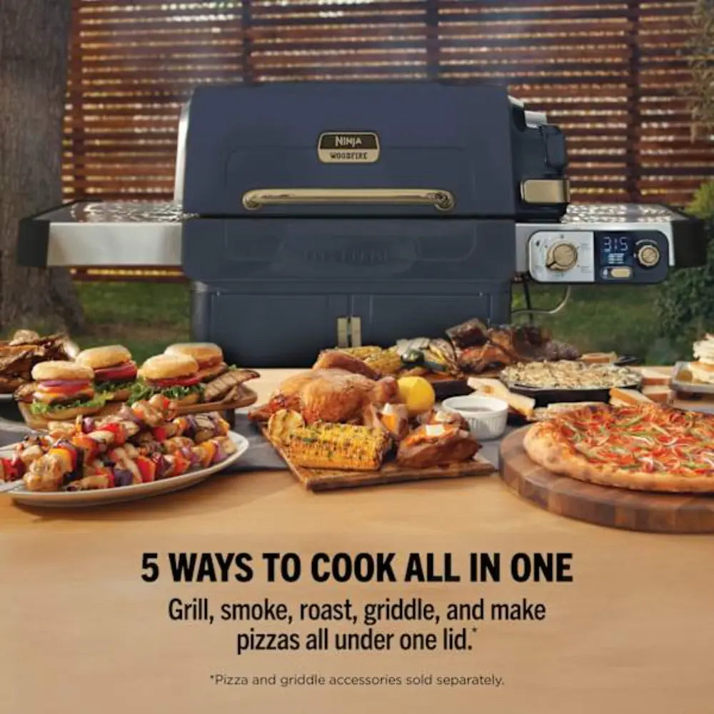 Ninja FlexFlame gas BBQ grill in navy blue with brass handles and digital control panel, displayed outdoors with various cooked foods including burgers, skewers, roasted chicken, corn, pizza and grilled items on wooden boards and plates in front, against a brick fence background