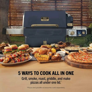 Ninja FlexFlame gas BBQ grill in navy blue with brass handles and digital control panel, displayed outdoors with various cooked foods including burgers, skewers, roasted chicken, corn, pizza and grilled items on wooden boards and plates in front, against a brick fence background
