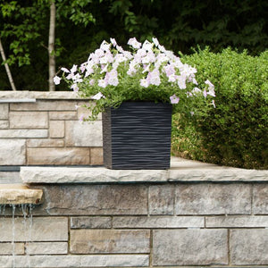Slate grey rippled garden planter with white and pale pink trailing petunias and green foliage, positioned on a natural stone wall with water feature and hedges in background