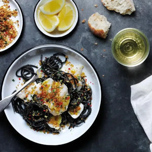 Overhead view of a white enamel dish containing black squid ink pasta with white fish, breadcrumb topping and red chili flakes, accompanied by lemon slices, white wine, crusty bread and seasoning on dark gray surface