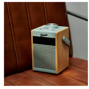 Roberts Rambler Midi DAB+ FM radio with Bluetooth in duck egg blue and natural wood finish, sitting on a brown leather surface with a burgundy leather chair in the background