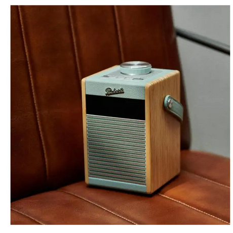 Roberts Rambler Midi DAB+ FM radio with Bluetooth in duck egg blue and natural wood finish, sitting on a brown leather surface with a burgundy leather chair in the background