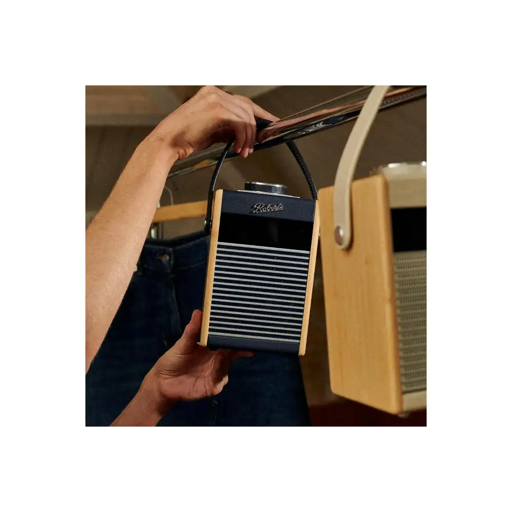 Person holding a Roberts Rambler Midi portable DAB+ FM radio with Bluetooth in navy blue and wood finish with horizontal grille design and shoulder strap, displayed against vintage cream-coloured radios in soft-focus background