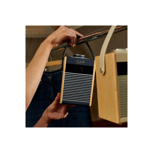 Person holding a Roberts Rambler Midi portable DAB+ FM radio with Bluetooth in navy blue and wood finish with horizontal grille design and shoulder strap, displayed against vintage cream-coloured radios in soft-focus background