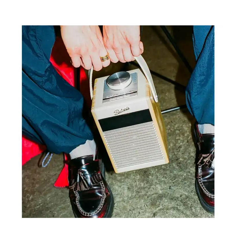 Roberts Rambler Midi DAB+ FM radio with Bluetooth in pastel cream being held by hands wearing blue jacket and black shoes, showing the device's speaker grille and dial interface