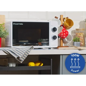 Russell Hobbs white manual microwave with 17 litre capacity and 700W power displayed on kitchen countertop next to striped tea towel, hanging kitchen utensils, and potted plants, with 700W power badge highlighted in blue circle