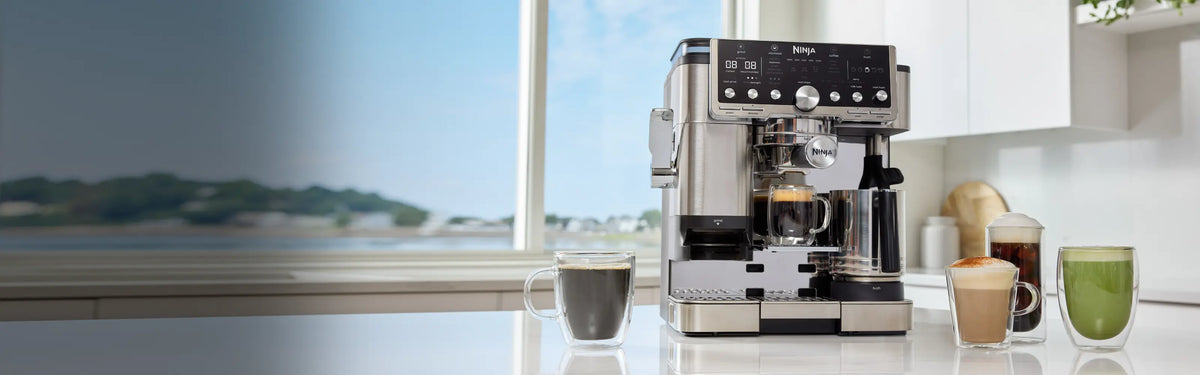 A sleek Breville espresso machine in stainless steel and black sits on a white countertop in a bright kitchen, with cups of coffee and latte beside it and a window view in the background.