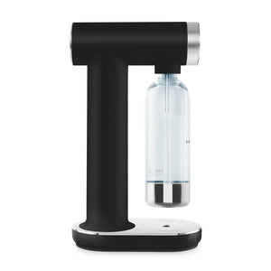 Smeg SKC01BLM matte black sparkling water maker with chrome accents, featuring a T-shaped design with carbonation canister attachment point and clear glass bottle with metal base mounted on white oval base