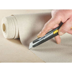 Hand holding Stanley snap-off utility knife with yellow and black handle cutting cream colored textured material, with roll of material and replacement blade pack visible in background