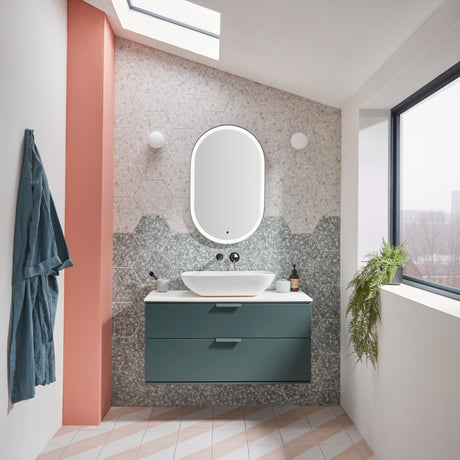 Modern bathroom vanity unit featuring a Juniper Green drawer cabinet with white countertop and vessel sink, paired with an oval illuminated mirror and grey mosaic tile backsplash in a contemporary spa-style setting with skylight and large window