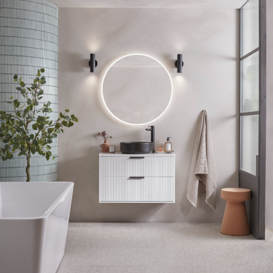 Modern white vanity unit with ribbed fronts and black handles beneath a circular illuminated mirror with black wall sconces in a minimalist bathroom setting with white freestanding bathtub, terracotta accessories and natural greenery