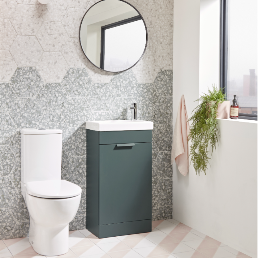 Modern bathroom vanity unit in Juniper Green with white ceramic sink and chrome tap, paired with white toilet, hexagonal grey mosaic tile splashback, round black framed mirror, and potted fern, on light grey and blush pink floor tiles