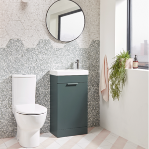 Modern bathroom vanity unit in Juniper Green with white ceramic sink and chrome tap, paired with white toilet, hexagonal grey mosaic tile splashback, round black framed mirror, and potted fern, on light grey and blush pink floor tiles