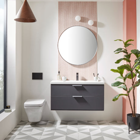 Modern bathroom vanity unit with Matt Carbon finish drawers and white countertop, paired with circular mirror against pink brick tile wall, white toilet, copper pendant light, geometric floor tiles, and potted green plant