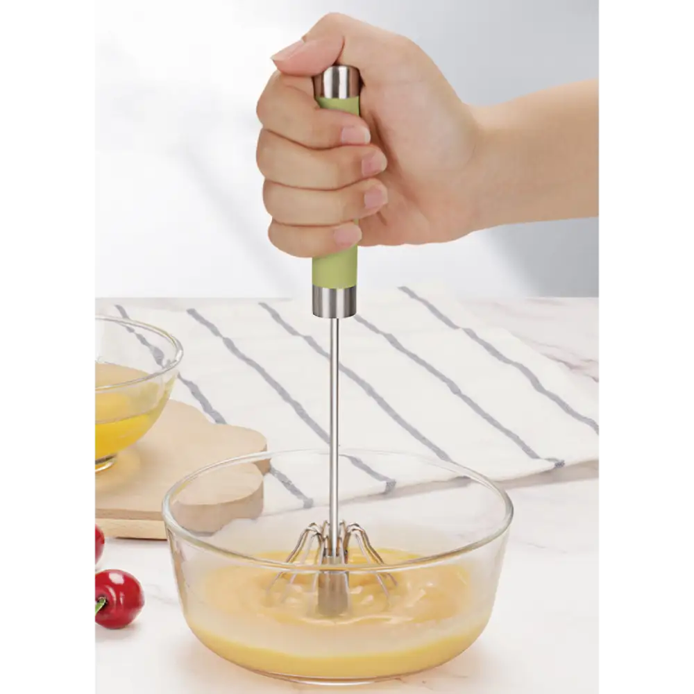 Child's hand holding a manual push spin whisk with green handle and stainless steel shaft, whisking pale yellow liquid in a clear glass bowl on a white striped kitchen towel, with fresh tomatoes and ingredients visible in background