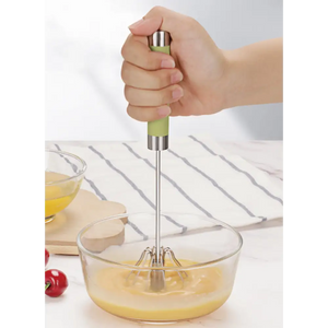 Child's hand holding a manual push spin whisk with green handle and stainless steel shaft, whisking pale yellow liquid in a clear glass bowl on a white striped kitchen towel, with fresh tomatoes and ingredients visible in background