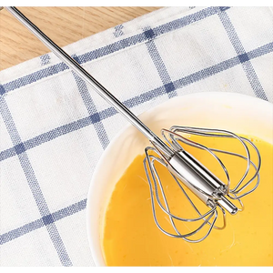 Manual push spin whisk with stainless steel handle resting in bowl of yellow liquid on blue and white checkered kitchen towel next to wooden floor