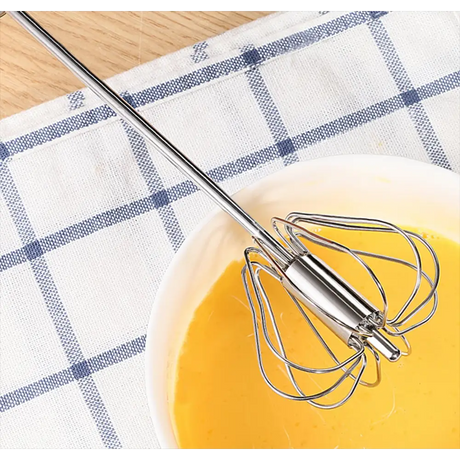 Manual push spin whisk with stainless steel handle resting in bowl of yellow liquid on blue and white checkered kitchen towel next to wooden floor