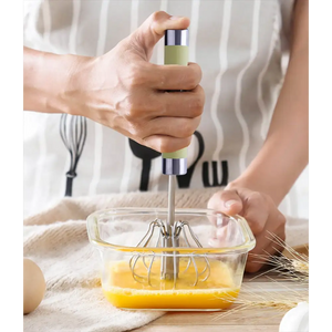 Hand using green and silver manual push spin whisk to mix yellow eggs in glass bowl on wooden surface with flour and wheat stalks, kitchen baking setup
