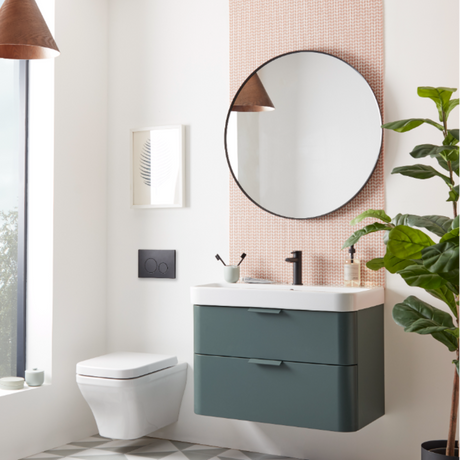 Modern bathroom interior featuring a Roper Rhodes System vanity unit in Derwent Blue with white countertop and black tap, circular black-framed mirror above, white wall-mounted toilet, pink brick-effect tiled accent wall, brown pendant lamp, potted green plant, and minimalist Scandinavian design aesthetic
