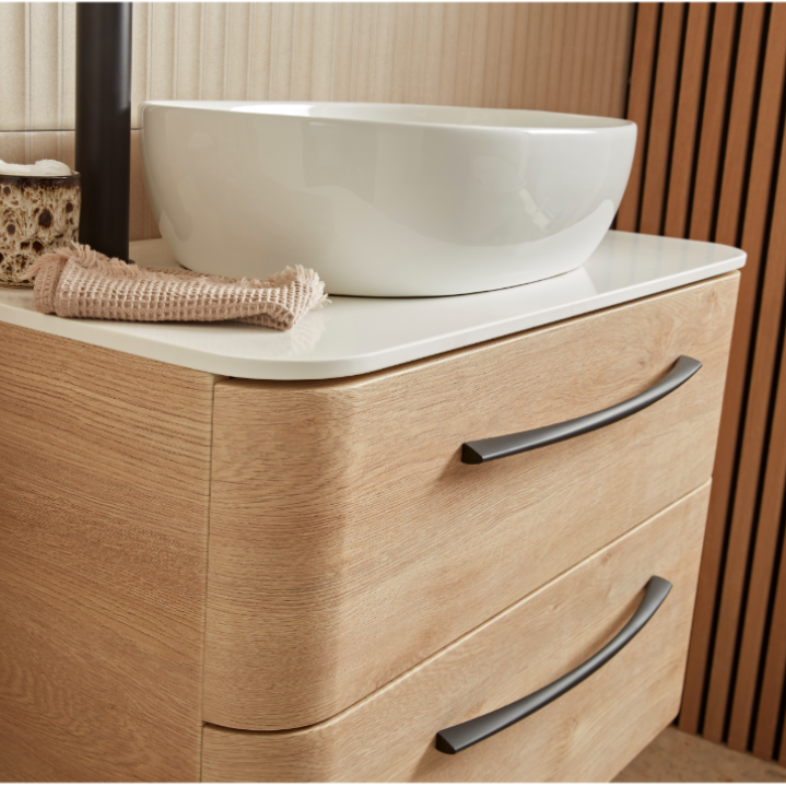 Sand Oak bathroom vanity unit with white ceramic vessel sink, curved white countertop, two dark metal handles, rolled towel and woven basket accessory, modern minimalist design with wood grain finish and warm beige background