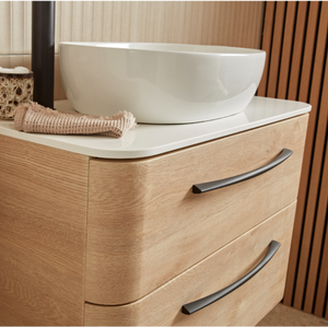 Sand Oak bathroom vanity unit with white ceramic vessel sink, curved white countertop, two dark metal handles, rolled towel and woven basket accessory, modern minimalist design with wood grain finish and warm beige background