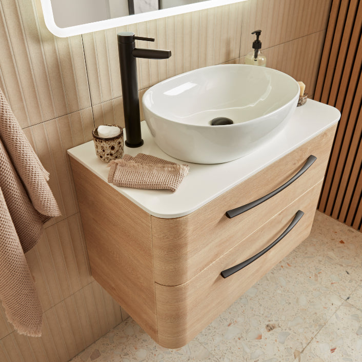 Modern light oak wood vanity unit with white countertop and oval white ceramic basin sink, black tall faucet, illuminated mirror, and matching beige towels in contemporary bathroom setting