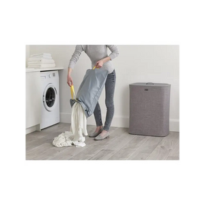 Woman emptying grey fabric laundry basket with yellow handles next to white washing machine and grey Joseph Joseph Tota 90 litre separation hamper on wooden floor