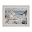Framed coastal scene painting showing whitewashed cottages overlooking a sandy beach with moored fishing boats, calm blue waters, and sailing yachts in the distance, in a cream-coloured wooden frame