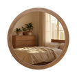 Round wooden wall mirror with natural oak frame reflecting a bedroom interior with bed, wooden dresser, potted plants and sunlit window