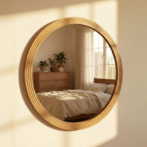 Large circular wooden wall mirror with natural oak frame featuring stepped detailing, reflecting a bright bedroom interior with wooden dresser, potted plants, bed with neutral bedding, and sunlit window with sheer curtains