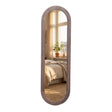 Tall pill-shaped standing mirror with natural wood frame reflecting bedroom interior with bed, window, potted plant and curtains