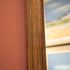 Tall pill-shaped dressing mirror with natural wood frame featuring vertical grain detail, positioned against coral pink wall with blurred landscape artwork visible in reflection
