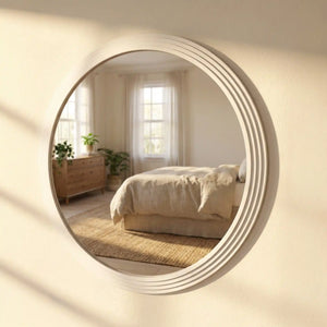 Large round oyster white wall mirror with stepped ribbed frame reflecting a minimalist bedroom interior with cream bedding, wooden dresser, potted plants and natural window light
