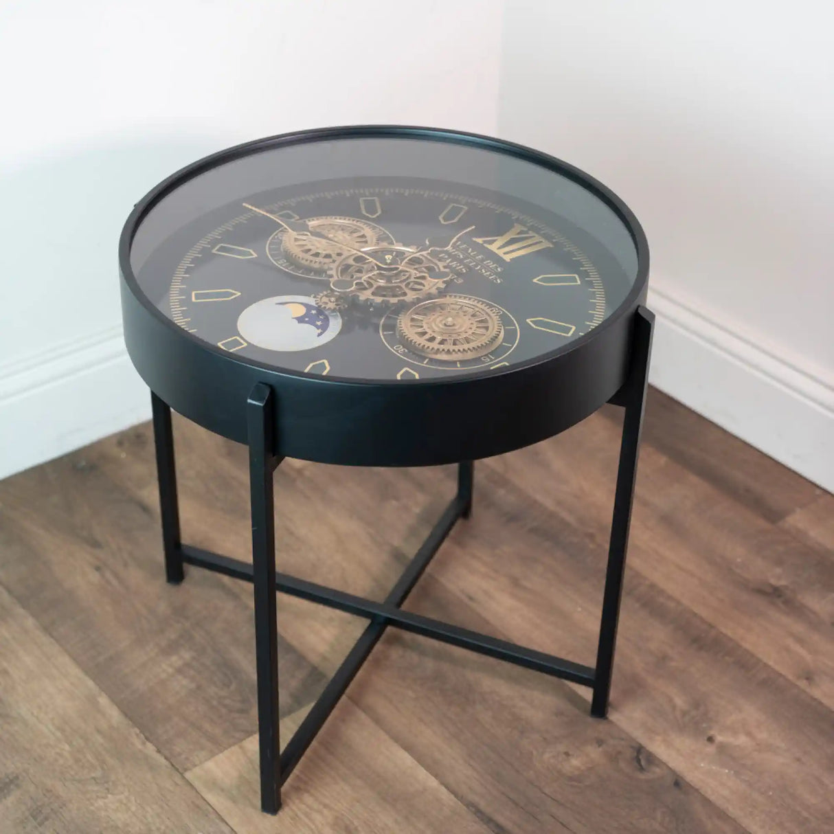 Black round side table with exposed gear clock face design, brass-colored gears and moon phase indicator on dark dial, metal frame with cross-base legs on wooden floor