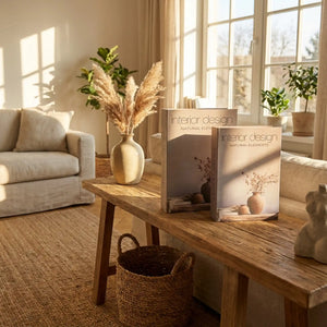 Two interior design coffee table books titled "Interior Design Natural Elements" displayed on a wooden table in a bright, minimalist living room with beige upholstery, pampas grass in a cream ceramic vase, potted green plants, woven basket, and natural sunlight from large windows