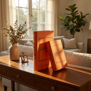 Two orange luxury book boxes displayed on a wooden console table beside a cream ceramic vase with dried eucalyptus, gold decorative object, and potted green plant, with a neutral sofa and windows in the background