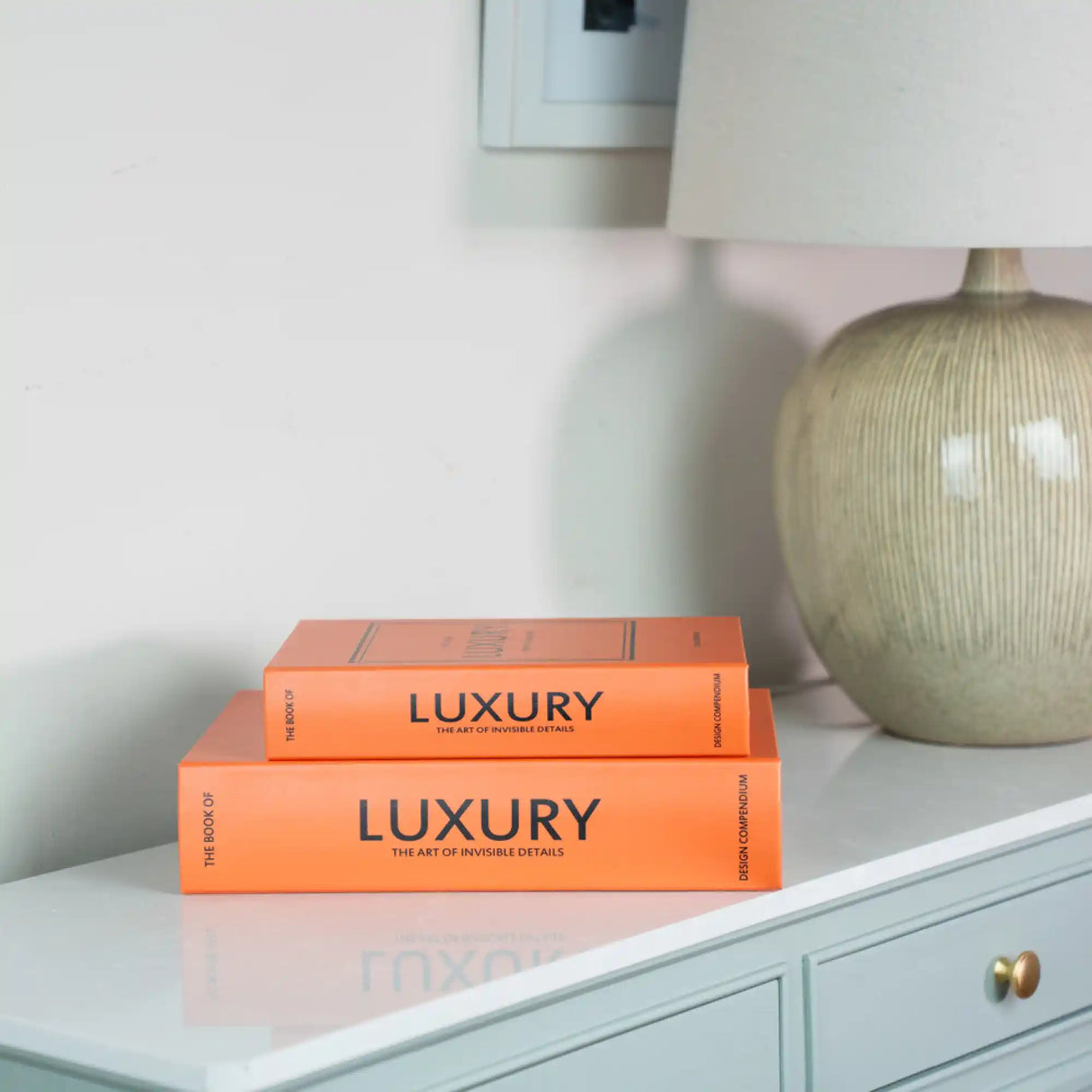 Two stacked orange decorative books titled "Luxury: The Art of Invisible Details" displayed on a white drawer unit next to a textured cream ceramic lamp base and a white thermostat on a pale wall