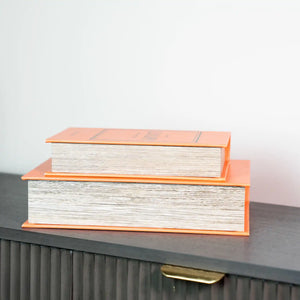 Two stacked decorative book boxes with bright orange covers and cream textured paper pages, displayed on a dark gray modern dresser with vertical ribbed detailing and gold hardware