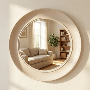 Large circular oyster-white framed mirror reflecting a minimalist living room with beige sofa, wooden bookshelf, and natural light from window, modern home decor accent piece