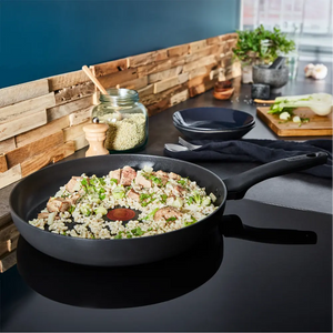 Tefal Ultimate 30cm black frying pan with cooked rice and meat dish on dark countertop beside wooden cutting board with vegetables and fresh herbs in modern kitchen setting