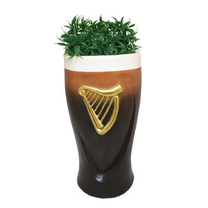 Black ceramic pint glass planter with white foam head rim and gold harp emblem, filled with green ornamental grass, Irish stout beer glass design