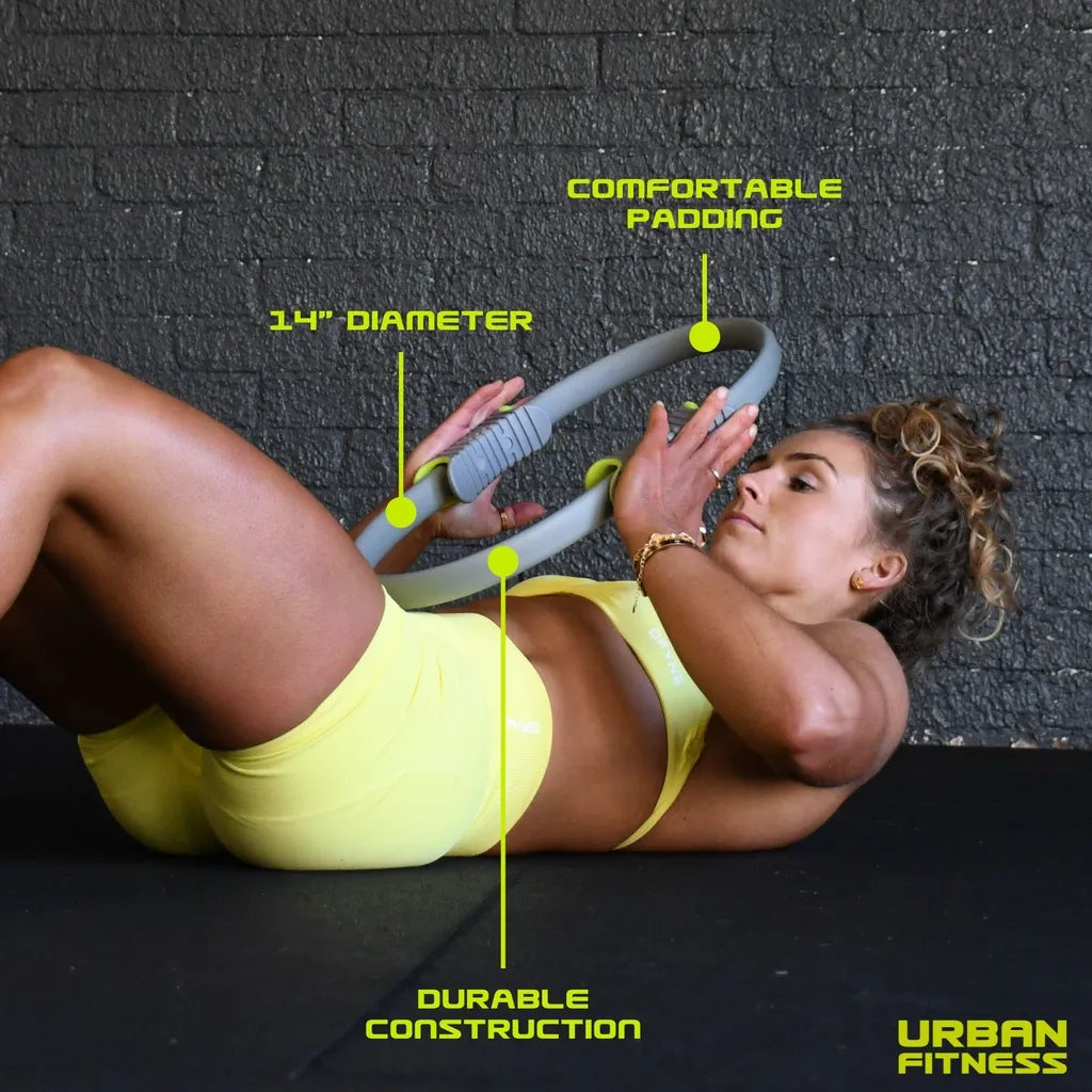 Woman in yellow sports bra and shorts performing pilates exercise with gray Urban Fitness 14 inch diameter pilates ring on black floor against brick wall, highlighting comfortable padding and durable construction features