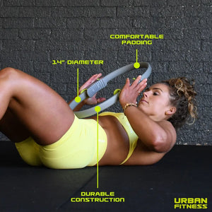 Woman in yellow sports bra and shorts performing pilates exercise with gray Urban Fitness 14 inch diameter pilates ring on black floor against brick wall, highlighting comfortable padding and durable construction features