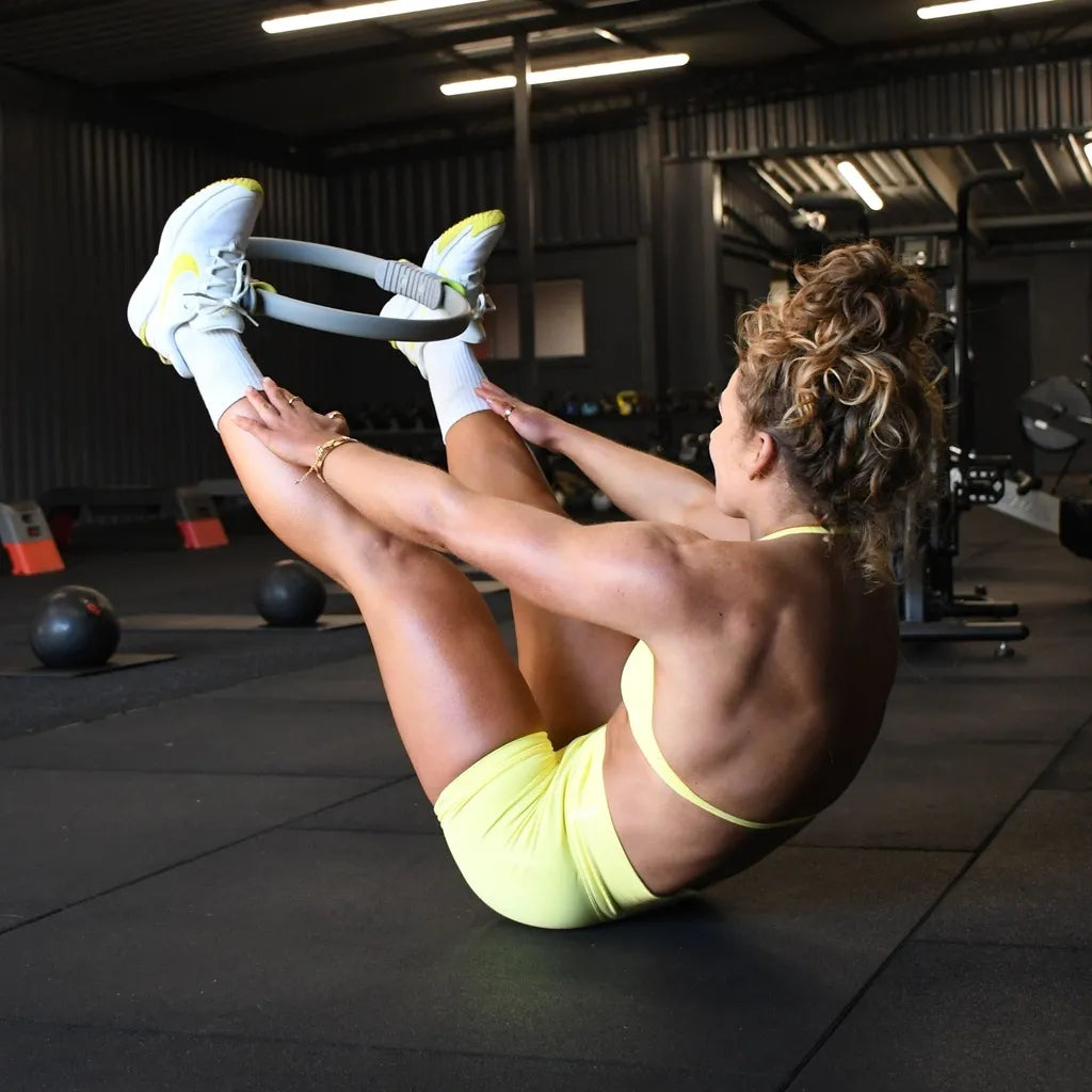 Woman performing a pilates exercise with a yellow and black resistance ring in a modern gym setting with white sneakers and neon yellow sports bra