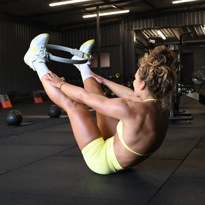 Woman performing a pilates exercise with a yellow and black resistance ring in a modern gym setting with white sneakers and neon yellow sports bra
