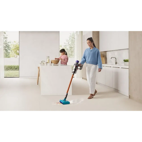 Woman using a Dyson Cyclone V10 Submarine cordless stick vacuum with red and teal base on light kitchen floor, child in pink in background at white kitchen island with modern minimalist design and garden views