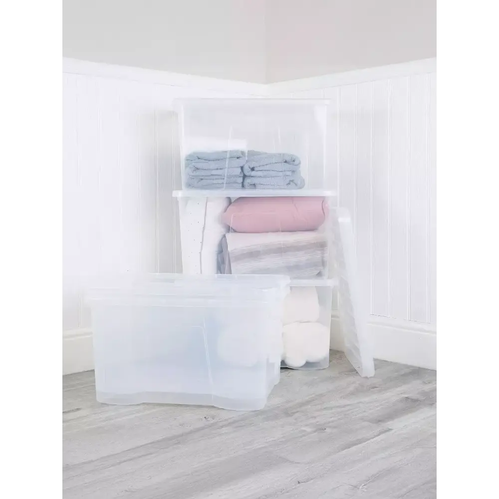 Clear plastic storage box with lid containing folded towels in blue, pink, and white, stacked on gray wooden flooring in a minimalist white bathroom setting