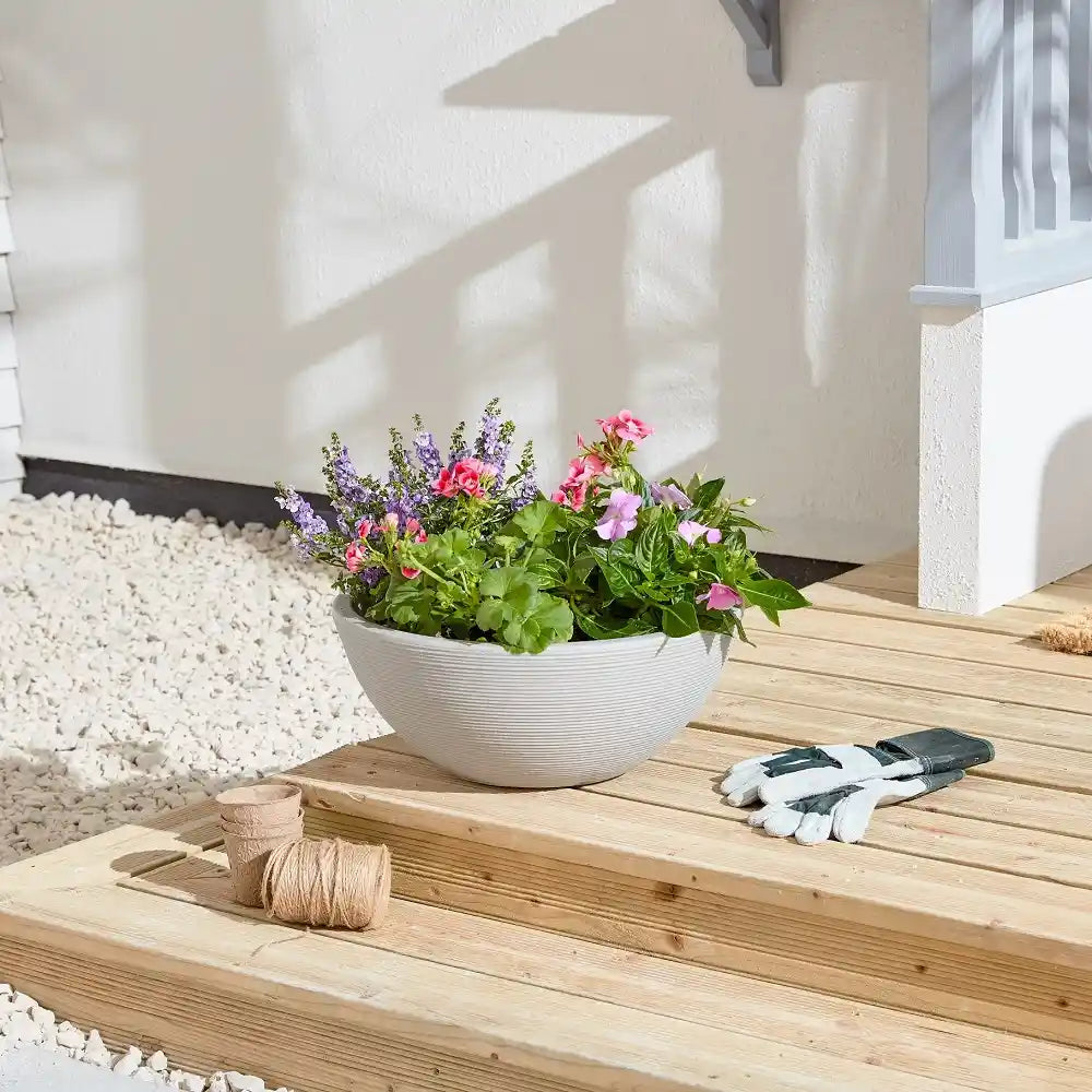 White ribbed ceramic garden planter with pink and purple flowering plants on wooden decking beside gardening gloves and twine, with white rendered house wall and gravel in background