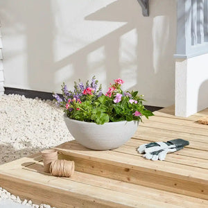 White ribbed ceramic garden planter with pink and purple flowering plants on wooden decking beside gardening gloves and twine, with white rendered house wall and gravel in background